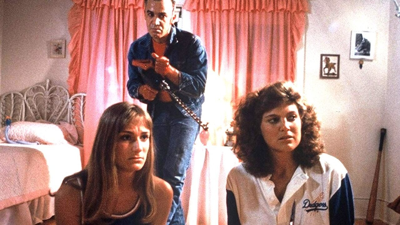 Two teen girls in pajamas sit on the floor in an overwhelmingly pink bedroom as a killer with a drill approaches silently in the background. From The Slumber Party Massacre (1982).