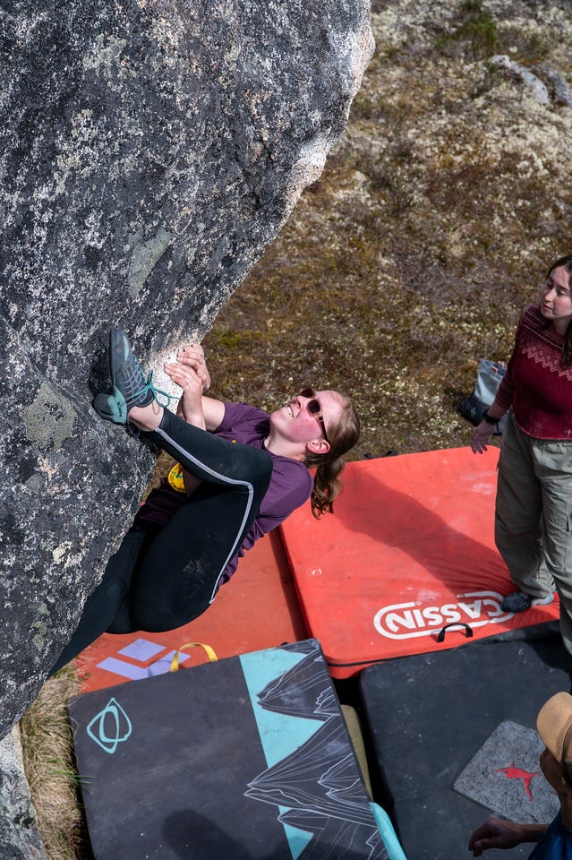 Shot from above: a girl in a purple shirt and dark leggings climbs the underside of a steeply overhanging boulder, body nearly horizontal, both feet hooked on a rail above her hands, smiling mid-move. Orange and patterned bouldering pads are stacked on the ground below. A spotter in a dark red sweater stands to the right watching. Shot from above: a girl in a purple shirt and dark leggings climbs the underside of a steeply overhanging boulder, body nearly horizontal, both feet hooked on a rail above her hands, smiling mid-move. Orange and patterned bouldering pads are stacked on the ground below. A spotter in a dark red sweater stands to the right watching.