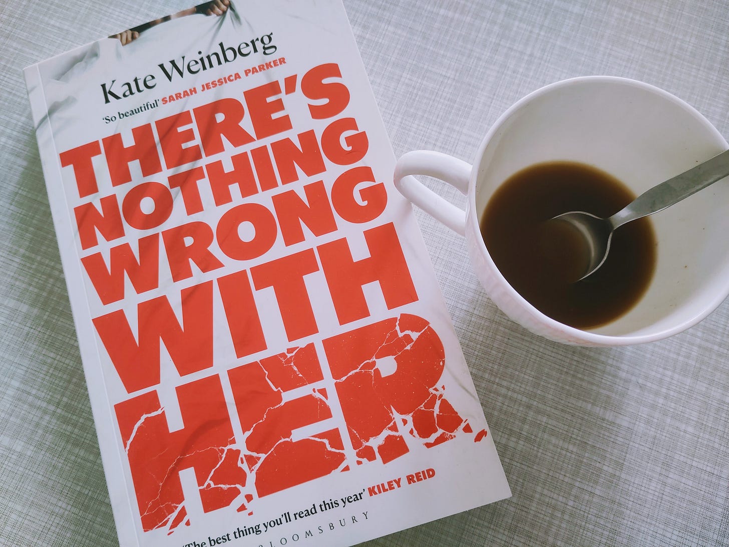 On a gray table top, there is a white, almost empty cup of coffee with a spoon, and Kate Weinberg's novel "There's nothing wrong with her". 