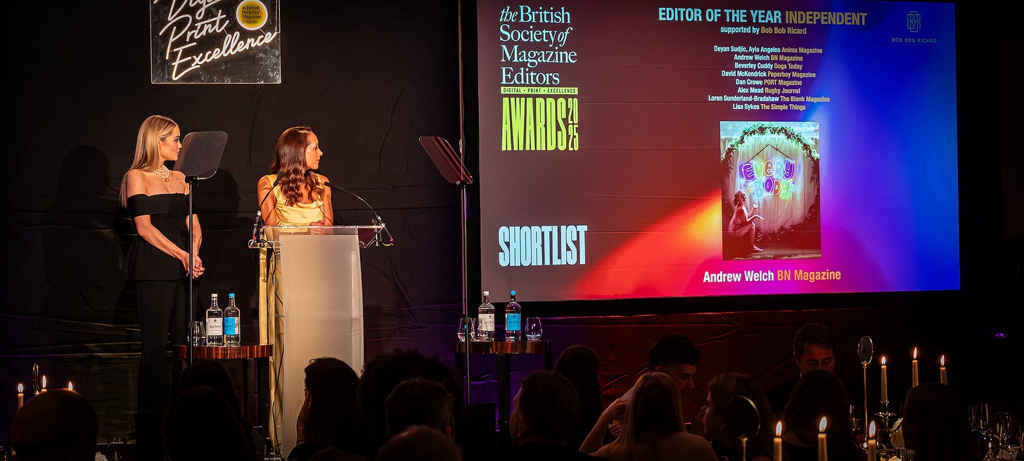 Two women stand at a podium on a candlelit stage at the 2025 BSME Awards. A large screen behind them displays the shortlist for Editor of the Year (Independent), including BN Magazine’s Andrew Welch, with a cover image of a nude woman seated in front of a neon sign reading “EveryBody.”