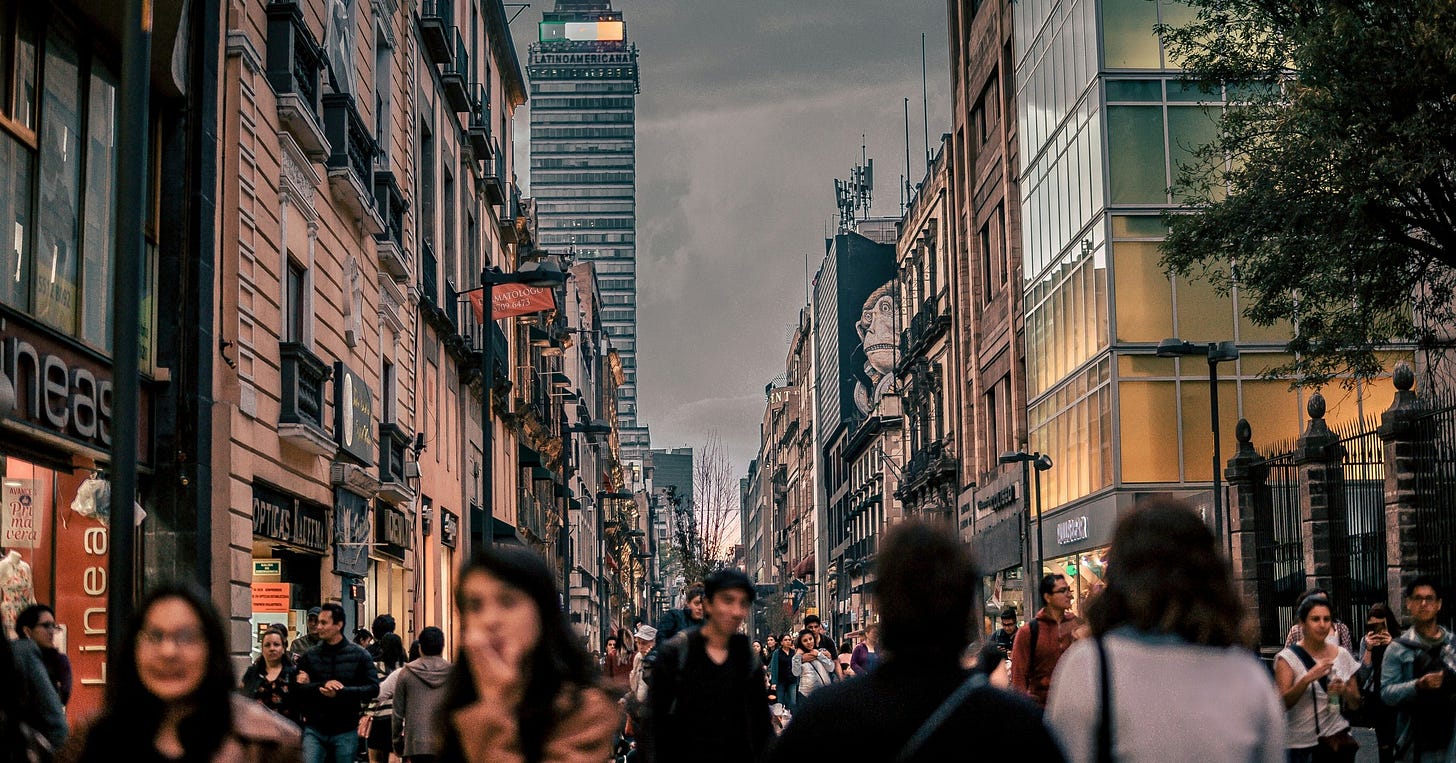 A crowded pedestrian street in Mexico City's historic centre at dusk, shopfronts on both sides and the Torre Latinoamericana rising at the end of the block.