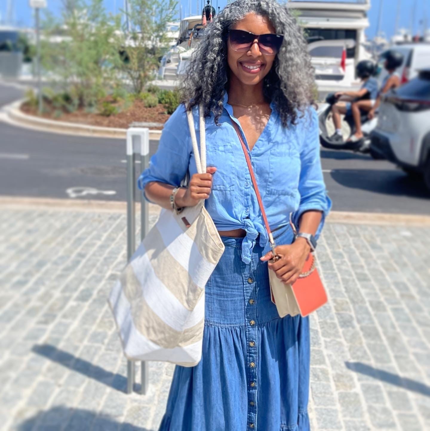 Khadijah wearing a blue linen top tied at the waist, a denim maxi skirt, sunglasses, and holding a striped straw beach bag smiling at the camera. Her hair is curly and flowing in the wind. Sailboats and motorcycles are in the background. It’s a sunny day with blue sky.