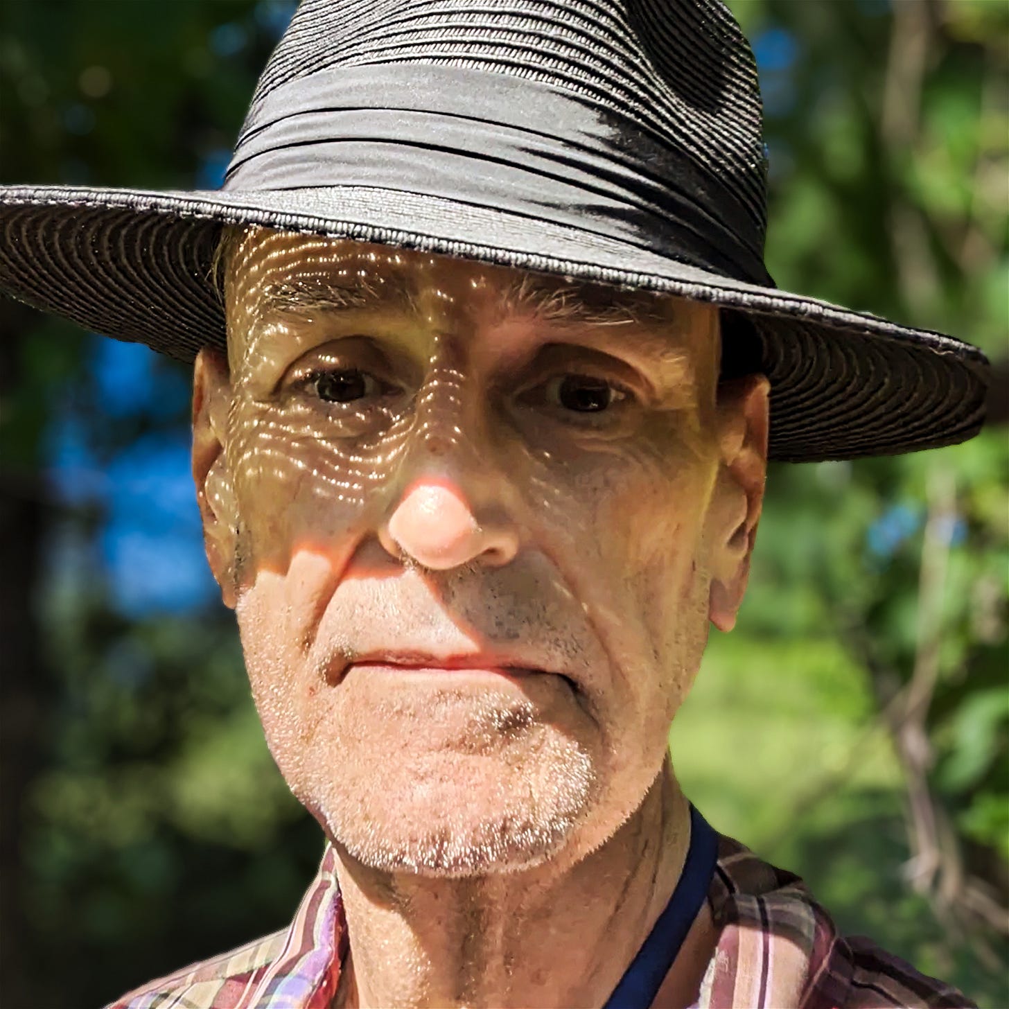 Johnny, 73, in his black, Panama hat. Dappled sunlight across his face. Plaid shirt. Indiana trees behind him. Looking straight at the camera.