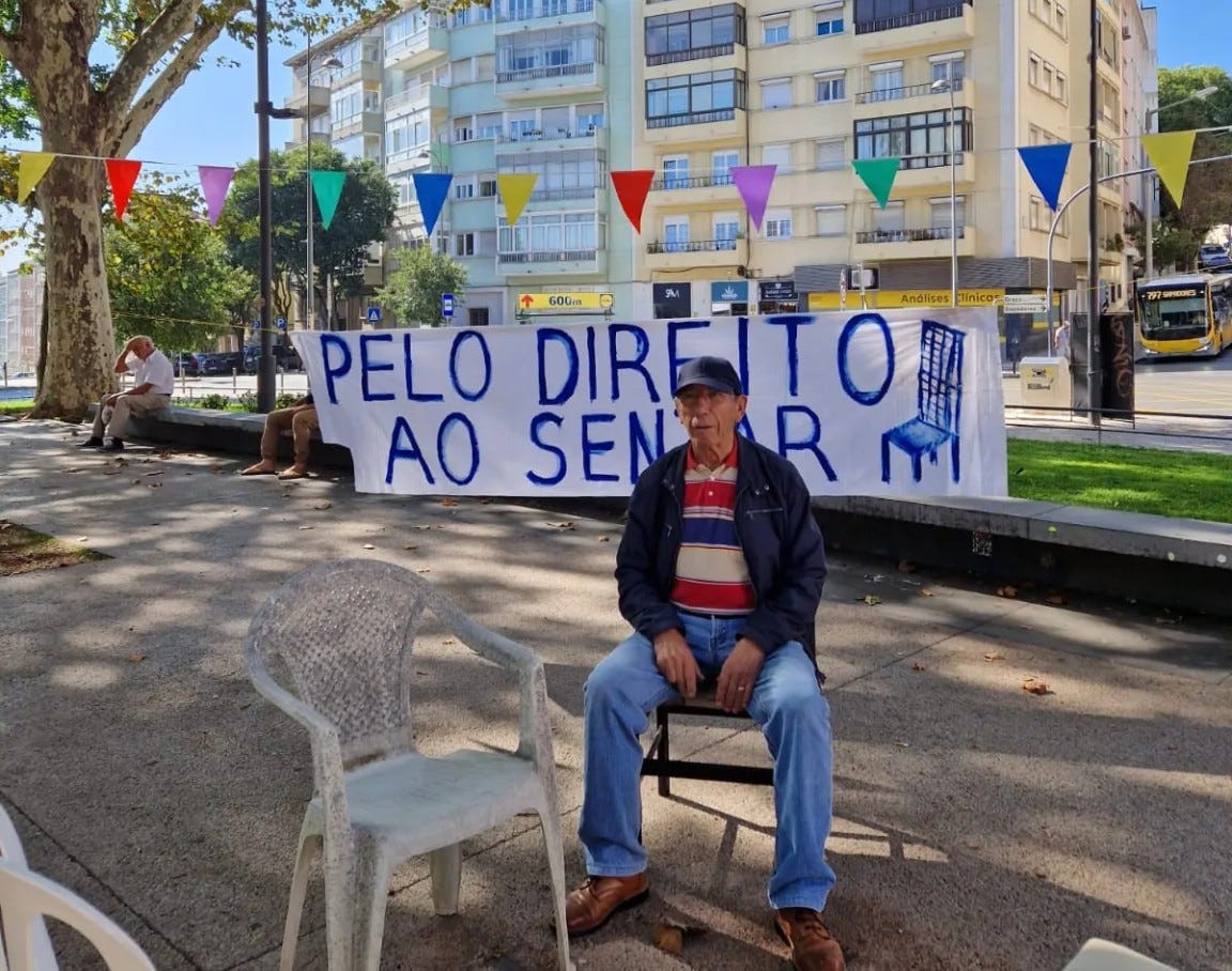 Un uomo anziano seduto su una sedia di metallo di fronte a uno striscione bianco con testo blu e un disegno di una sedia blu. Lo striscione recita "PELO DIREITO AO SENAR" (PER IL DIRITTO A SEDERSI). Dietro di lui, ci sono alberi, un edificio color crema con balconi e bandierine triangolari colorate appese tra i pali della luce. La scena si svolge all'aperto in una giornata luminosa.