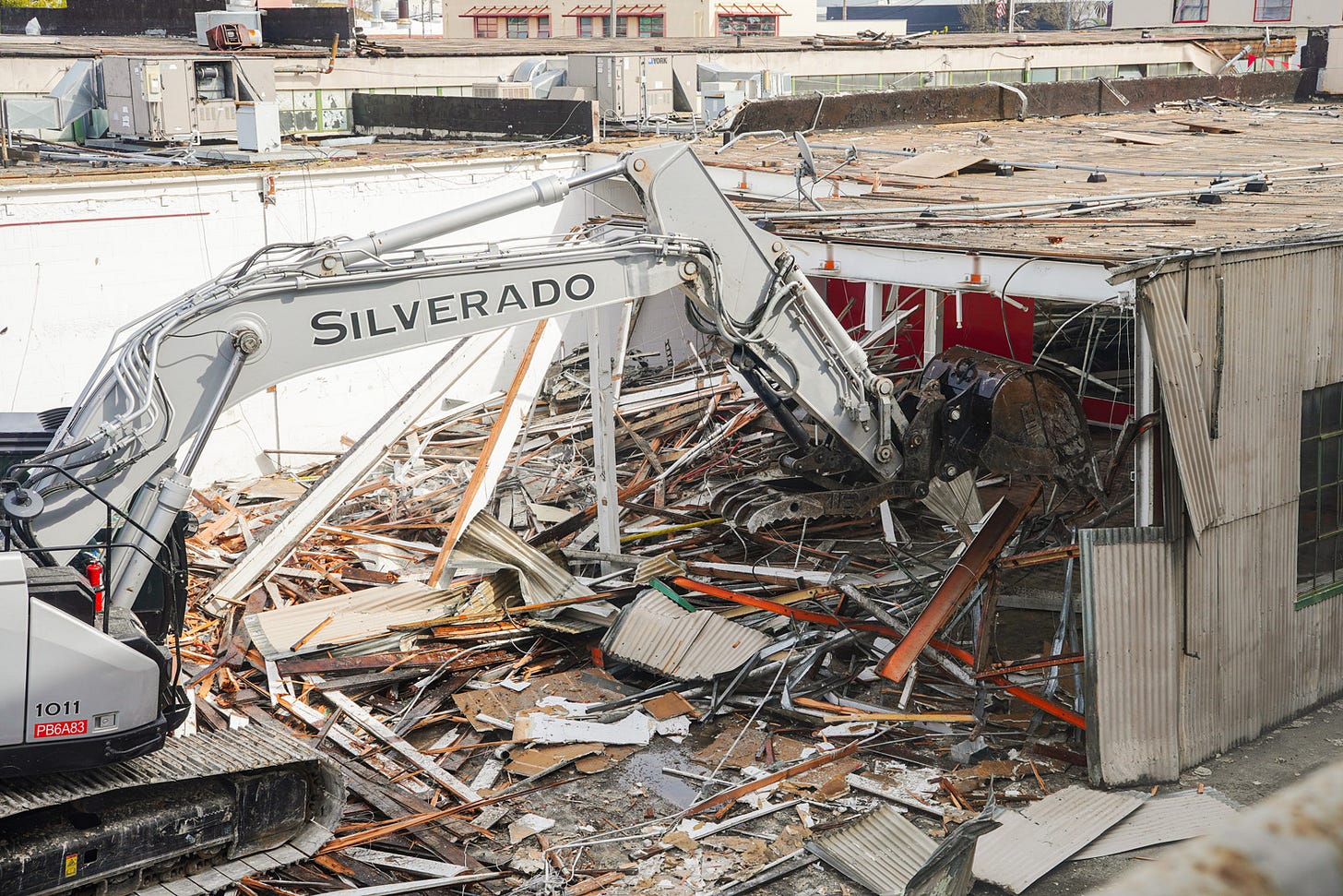 A large Silverado-branded excavator is actively demolishing a corrugated metal building, using its claw attachment to tear through the structure’s walls and roof. Piles of debris, including wooden beams, metal panels, insulation, and shattered materials, cover the ground around the demolition site. The building’s red interior walls and white support beams are partially exposed as the structure collapses. In the background, rooftop HVAC units and neighboring commercial buildings are visible, indicating an urban or industrial setting. The scene captures active construction demolition, heavy machinery, and structural dismantling.