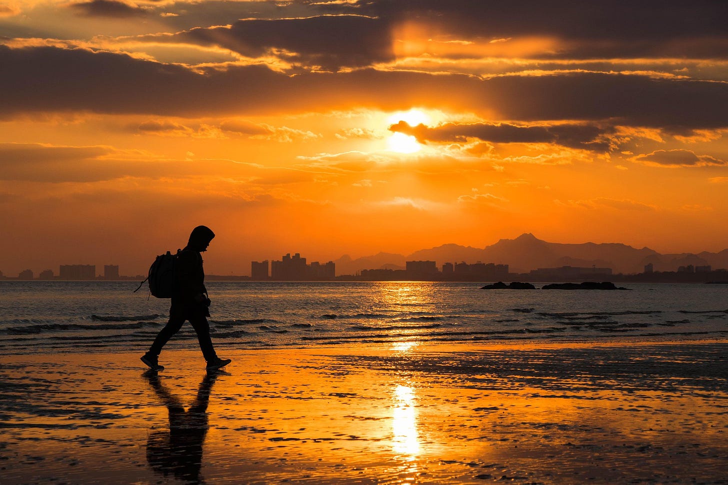 Fotografia em cores de pôr do sol em praia em Beidaihe. Em primeiro plano, há o perfil de uma pessoa caminhando. Ao fundo, é possível ver algumas construções.