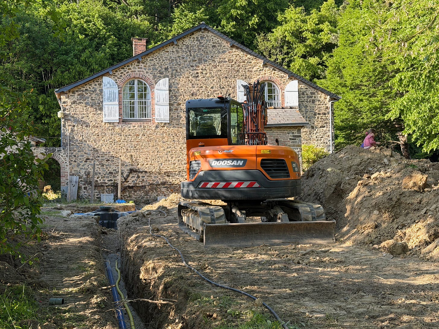An orange Doosan mini digger parked beside a long, freshly dug trench with pipes laid in it, in front of a stone coach house with arched windows and white shutters, surrounded by green trees in late afternoon light. An orange Doosan mini digger parked beside a long, freshly dug trench with pipes laid in it, in front of a stone coach house with arched windows and white shutters, surrounded by green trees in late afternoon light.