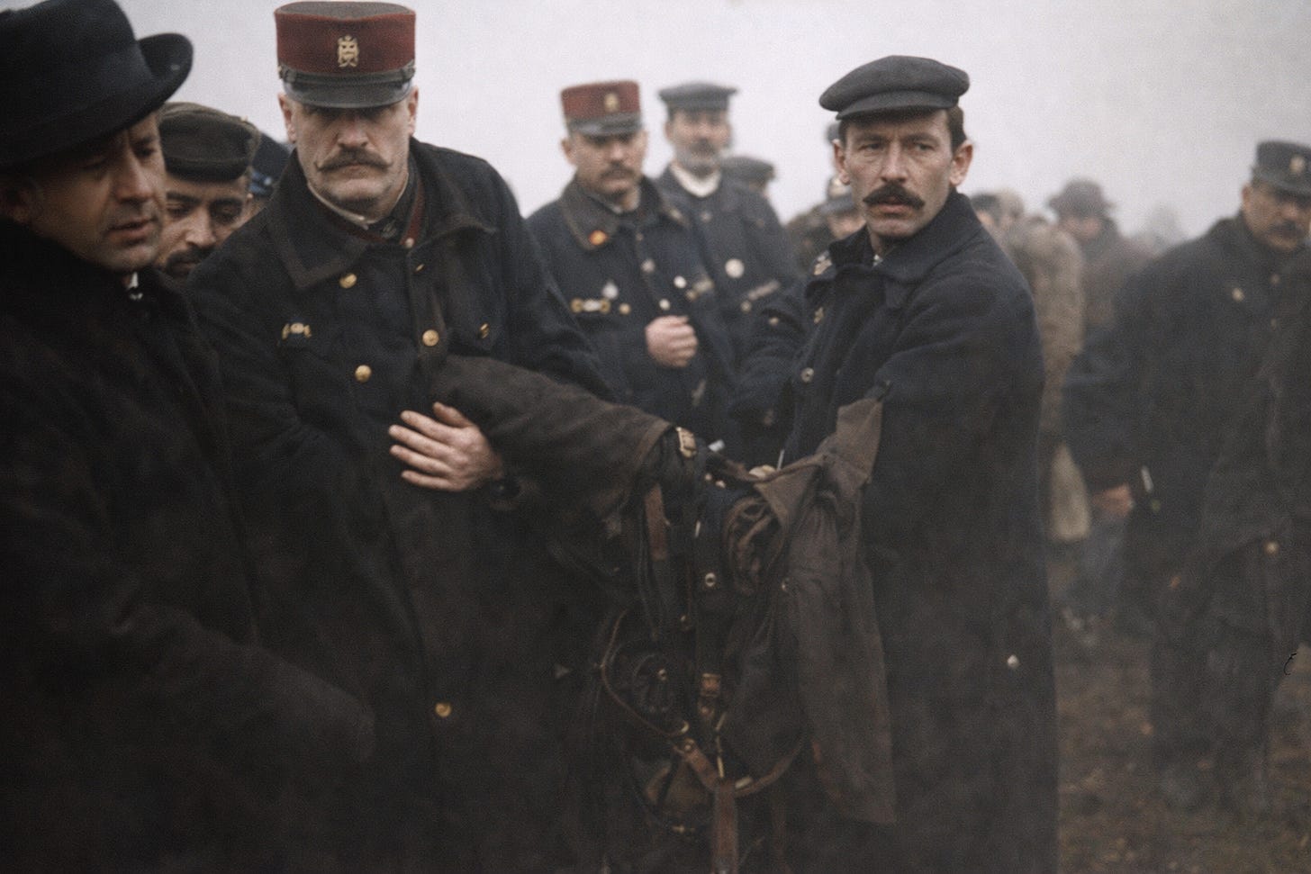 This scene shows a group of early 20th-century men—likely police officers and officials—gathered at the site after Franz Reichelt’s fatal jump. They are dressed in heavy coats and period hats, standing close together in a somber, foggy outdoor setting. At the center, a few men appear to be handling or carrying part of the parachute suit or related equipment, their expressions serious and focused. Others stand behind them, watching quietly. The mood is tense and subdued, reflecting the aftermath of a tragic event, with attention fixed on what has just occurred rather than on the camera.