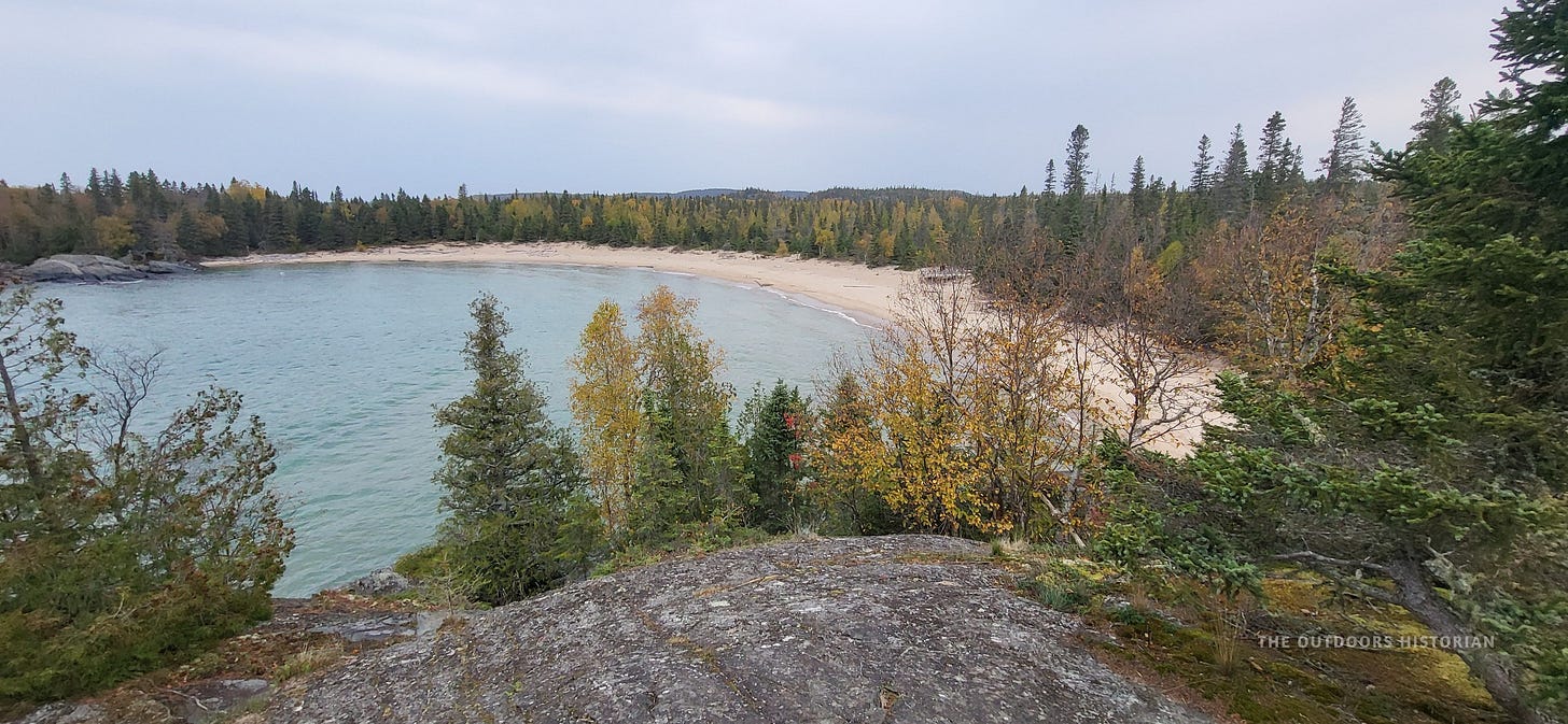 An elevated view looking down at a curved, sandy beach nestled among a dense pine forest, with the calm, protected waters of Lake Superior in the background.