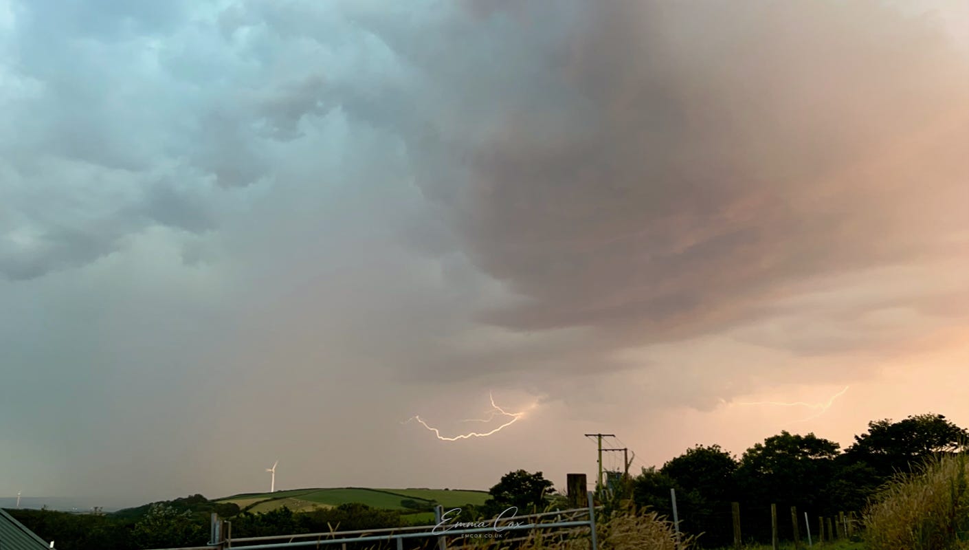 a moody orange/blue dusk sky with fork lightning. The view looks over countryside and a wind turbine before the landscape is lost in the distant rain.
