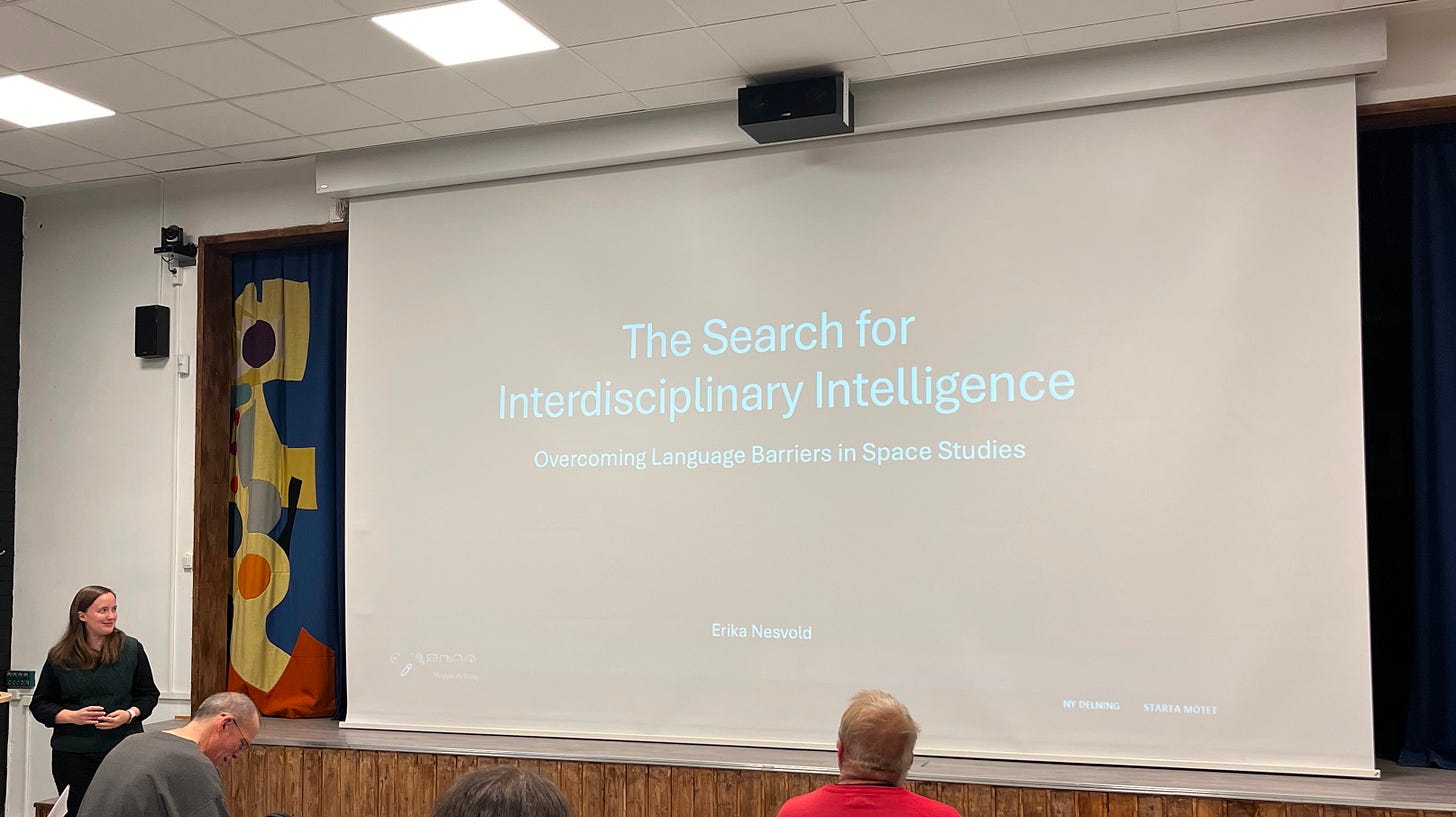 A photo of Erika Nesvold standing in front of a projector displaying the title slide of her talk, with text that reads "The Search for Interdisciplinary Intelligence: Overcoming Language Barriers in Space Studies" A photo of Erika Nesvold standing in front of a projector displaying the title slide of her talk, with text that reads "The Search for Interdisciplinary Intelligence: Overcoming Language Barriers in Space Studies"