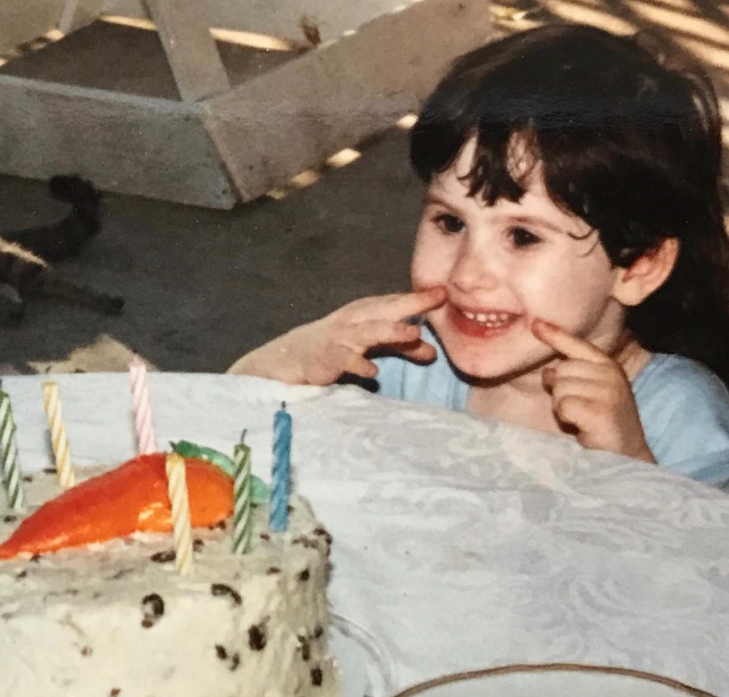 Little Lyric, holding their fingers to their face, making a “smile” for the photo as a carrot cake, a cake they already know they don’t like, is about to be served to them. Little Lyric, holding their fingers to their face, making a “smile” for the photo as a carrot cake, a cake they already know they don’t like, is about to be served to them.
