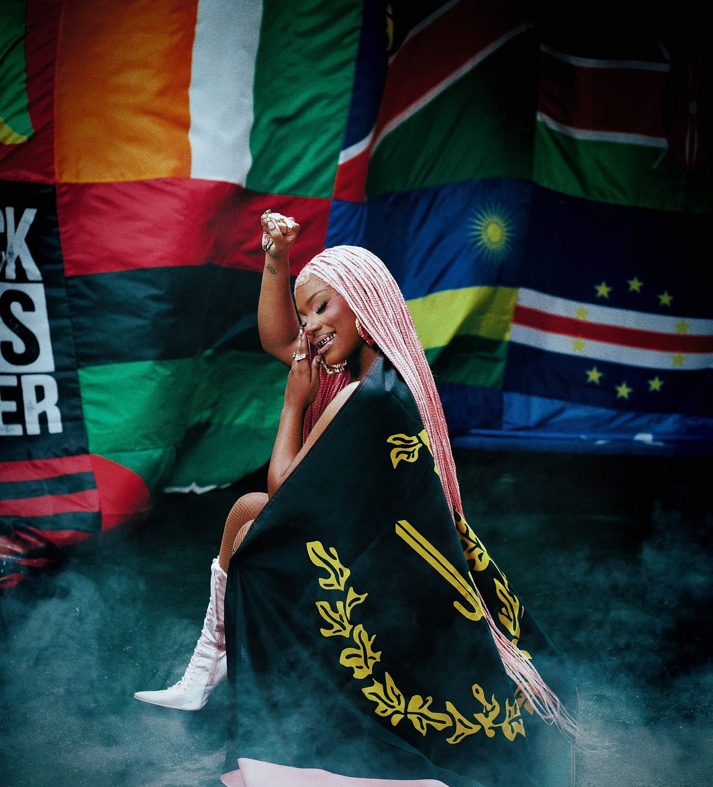 Rapper Monaleo kneeling with a fist raised, draped with the Black American Heritage flag, with other Black diaspora flags in the background.