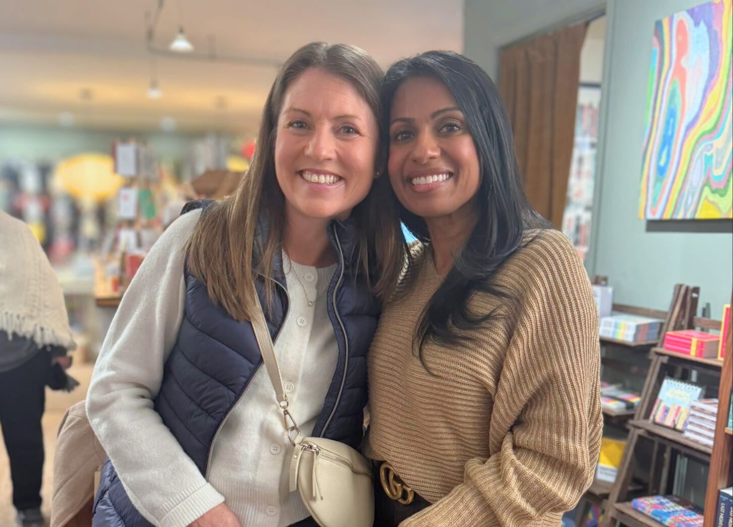 Amy Julia and Niro stand close together in a cozy bookshop, smiling at the camera. Colorful artwork and shelves of books are in the background.