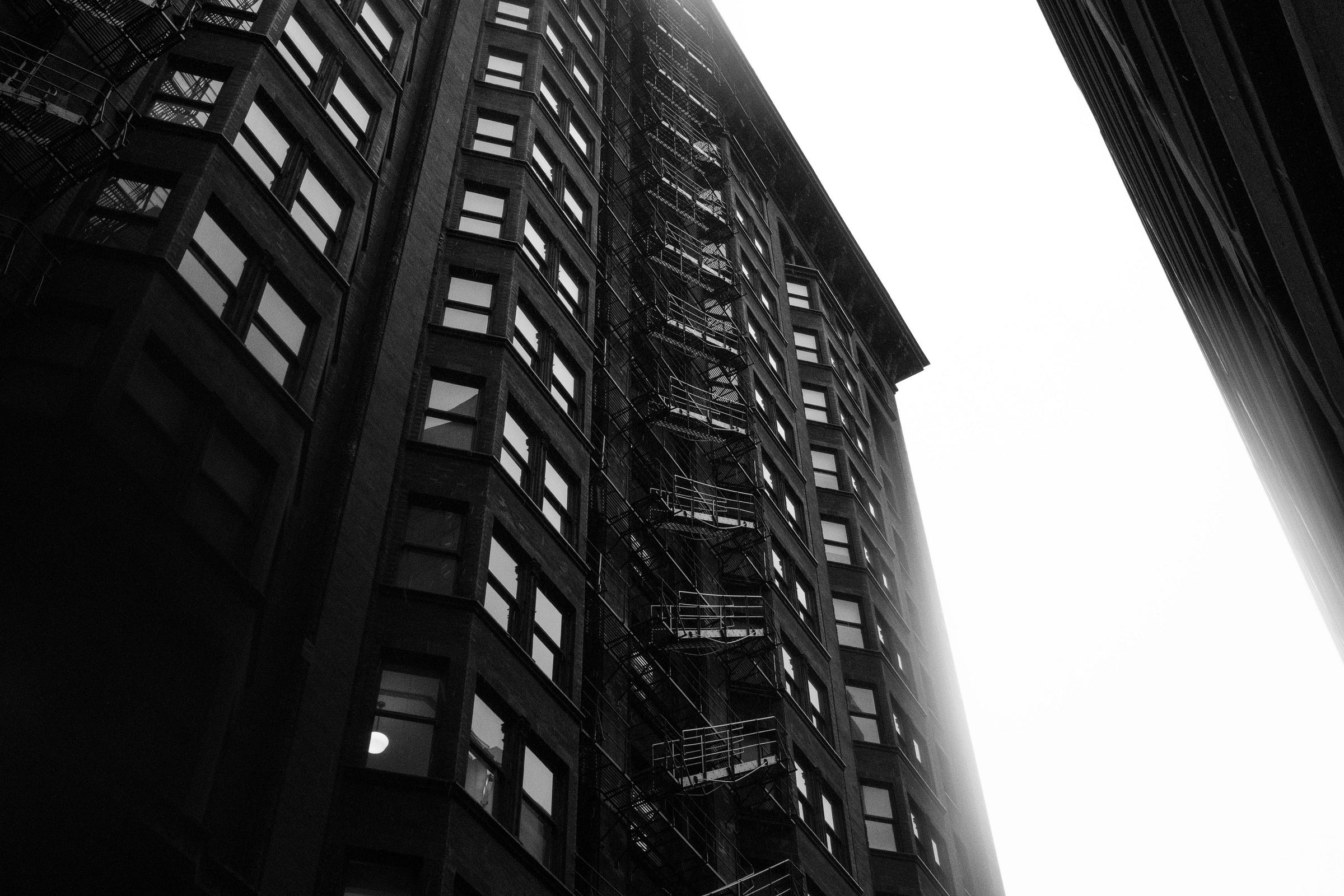 A towering historic building with a fire escape zigzagging up its façade, captured from a low angle against a bright, overcast sky. A towering historic building with a fire escape zigzagging up its façade, captured from a low angle against a bright, overcast sky.