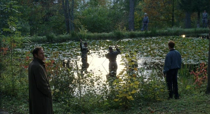 Several uniformed searchers wade through a pond surrounded by thick foliage, while two men watch from the edge of the water. The image hints at a search or investigation unfolding in a remote location.