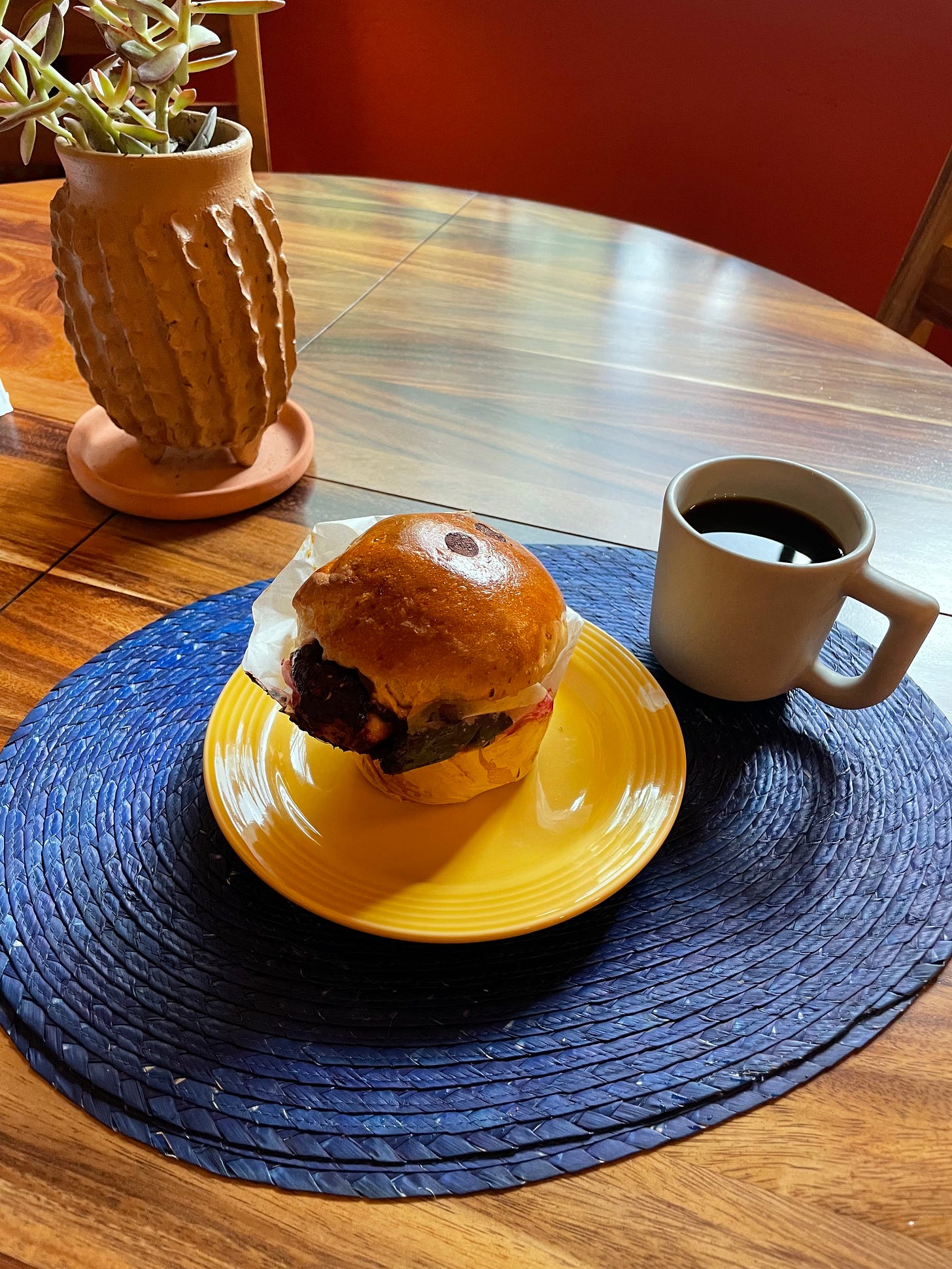 A glotón sitting on a plate next to a cup of coffee. The coffee and plate are on a round blue placemat on a wooden table, with a potted succulent on the left side of the frame.