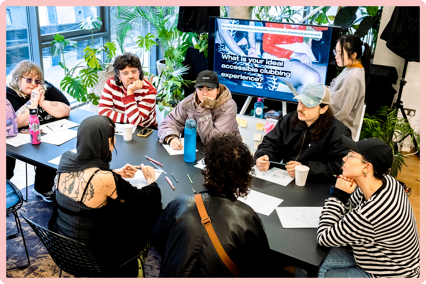 A photo of eight young disabled people sat around a table. Several of them are resting their head in their hands, in thought. They are also holding pens or coloured pencils, and paper. Behind them, text on a screen reads "What is your dream accessible clubbing experience?".