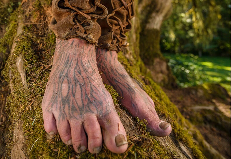 Close-up photograph of tattooed bare feet standing on moss-covered ground in a forest setting, with earthen hiking boots visible above the ankles.