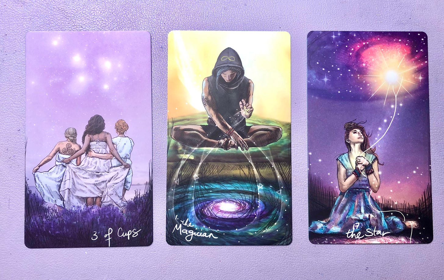 Three tarot cards arranged in a row: Three of Cups showing three figures in white robes against a purple starry background, The Magician card featuring a hooded figure in meditation pose above a cosmic vortex, and The Star card with a woman in flowing dress gazing up at a brilliant star in a purple night sky.