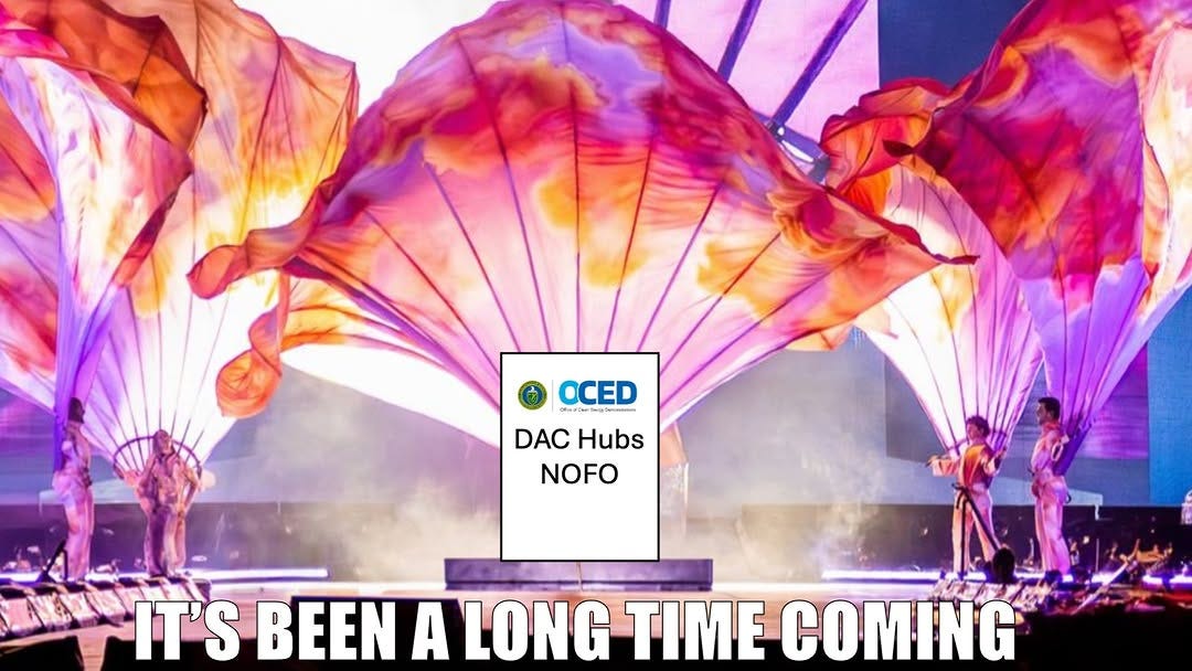 A stage performance with huge billowing fabric wings around a sign reading "OCED DAC Hubs NOFO", captioned "It's been a long time coming".
