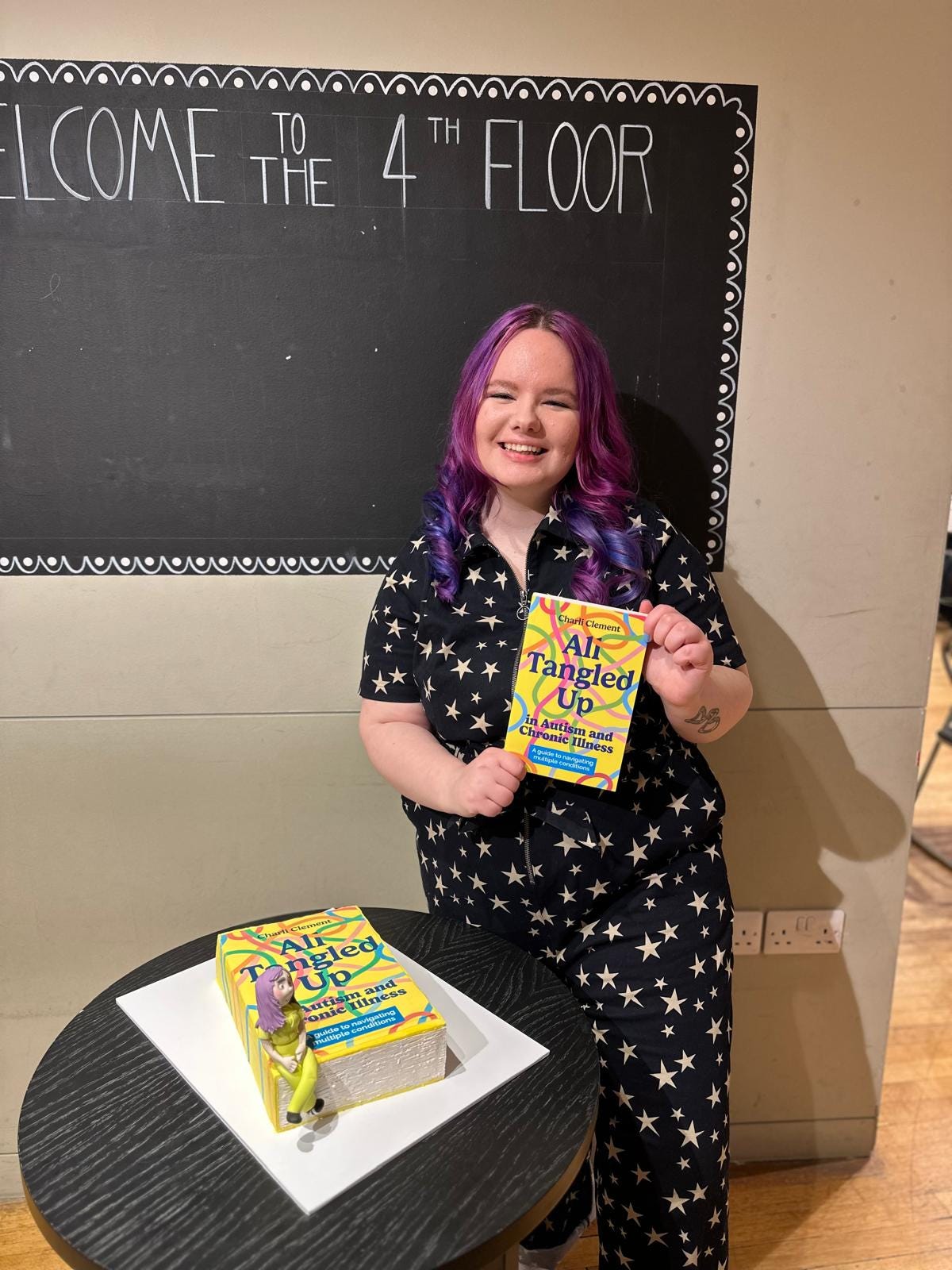 Charli wears a starry jumpsuit and beams at the camera holding their book. Next to them on a table is a cake made to look like the book with a little fondant version of them sat atop it