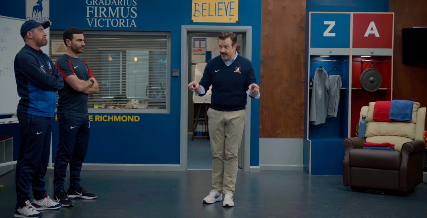 Scene from the TV show Ted Lasso showing Coach Ted Lasso standing in a locker room between his assistant coaches, Beard and Roy Kent. Ted is gesturing with both hands while talking, with a yellow “BELIEVE” sign hanging above the doorway behind him. The locker room walls are blue, with lockers labeled “Z” and “A” and a beige recliner chair with folded red and blue towels nearby. Scene from the TV show Ted Lasso showing Coach Ted Lasso standing in a locker room between his assistant coaches, Beard and Roy Kent. Ted is gesturing with both hands while talking, with a yellow “BELIEVE” sign hanging above the doorway behind him. The locker room walls are blue, with lockers labeled “Z” and “A” and a beige recliner chair with folded red and blue towels nearby.
