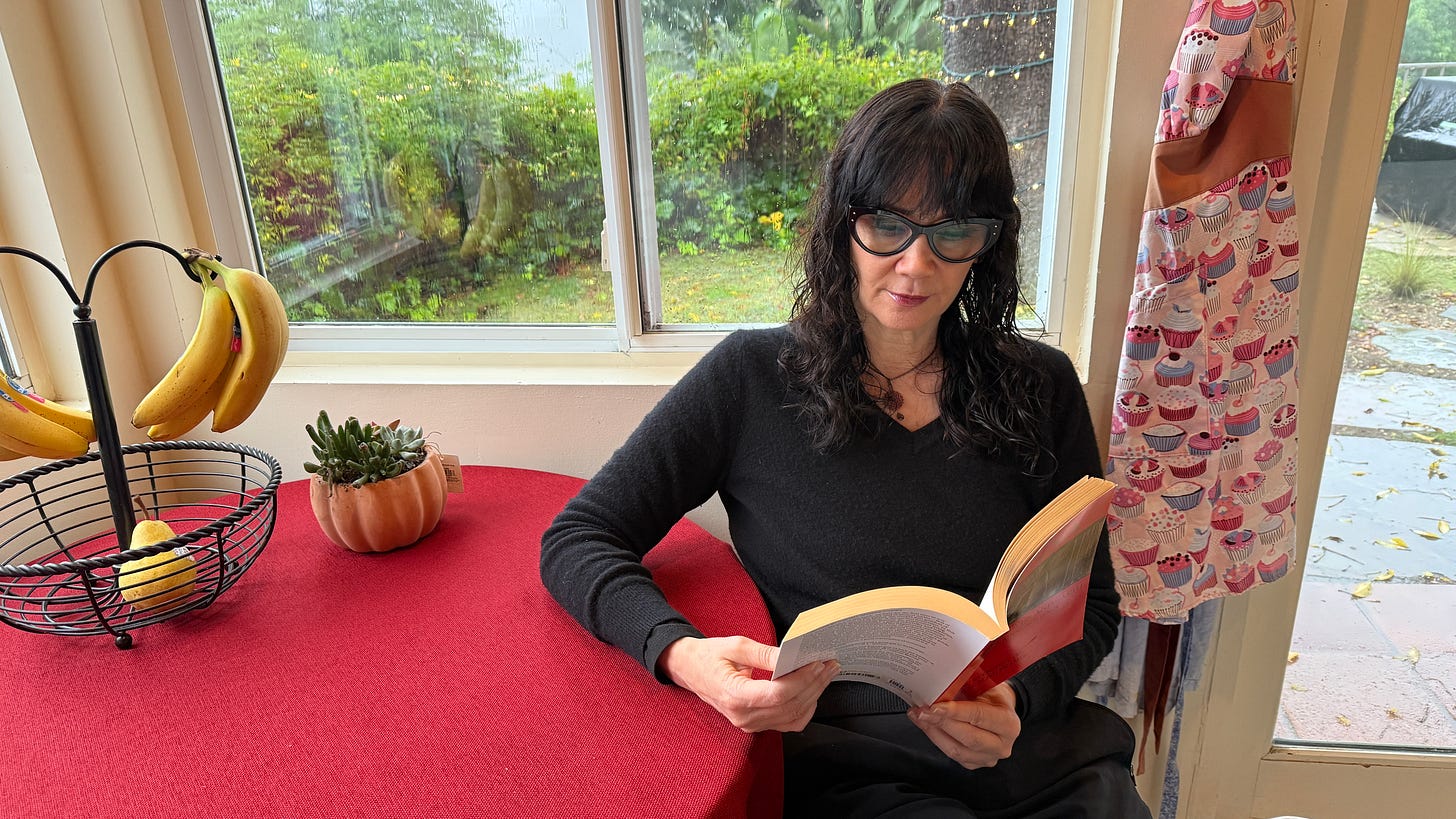 Laverne sitting at a small dining table with a red cloth cover while reading a book. There are bananas hanging and a pear in the basket on top of the table alongside a potted succulent plant. She is surrounded by windows and the tone is calm.