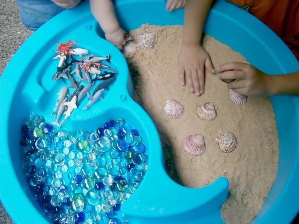 2 photos of play sand with small hands playing in them. The first features a bright blue plastic tray shaped into a circle with raised barriers in the middle for separating the plastic shark toys, the blue marbles and the sand. The second image features bright pink sand shaped into a star with cookie cutters.