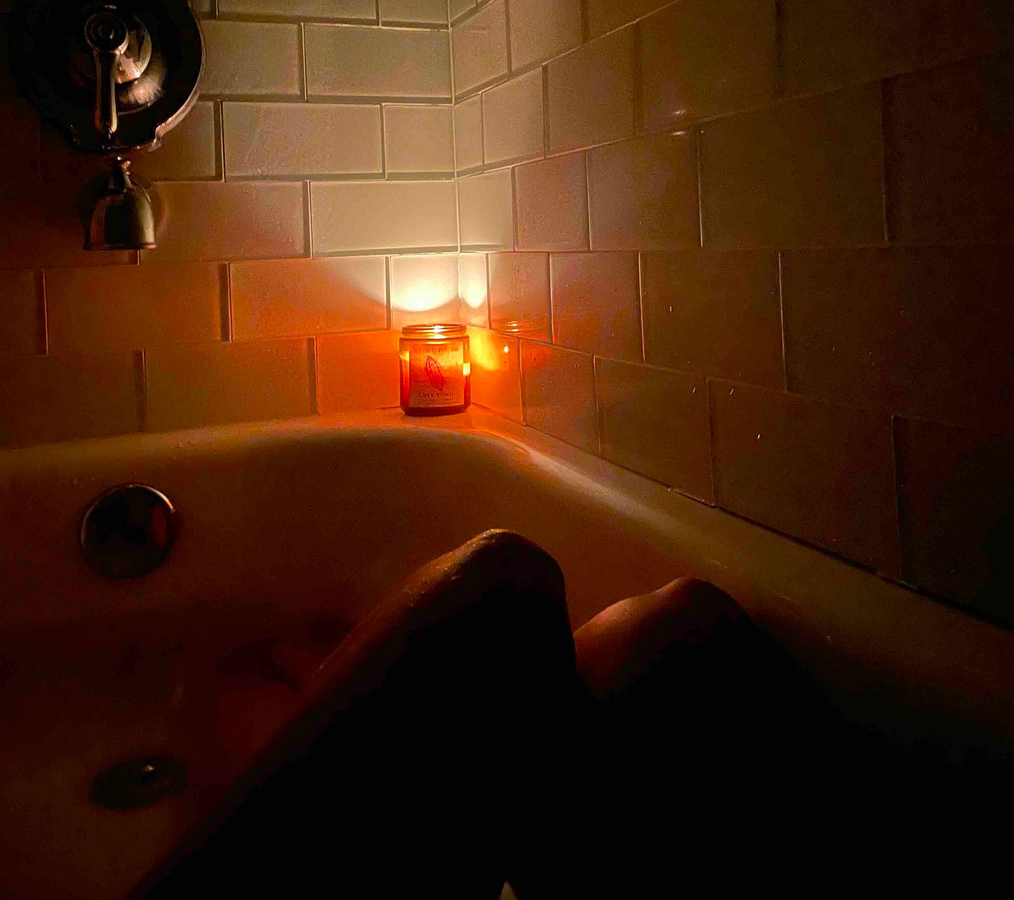 Image depicts a candle burning in the corner of an otherwise dark bathtub with knees leaning to the right