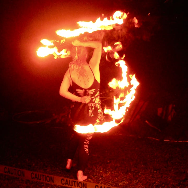 Two low-light photos of a fire performer spinning flaming props during MoonGroove's Saturday night show, trails of fire glowing orange and red against the dark sky.