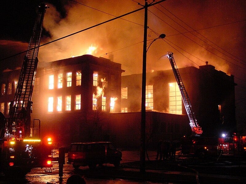 color photo of a century-old high school building fully engulfed in flames against a night sky. A tiny stream of water from a hose at the top of a truck's tall ladder is dwarfed by the conflagration, only emphasizing futility of trying to douse the fire color photo of a century-old high school building fully engulfed in flames against a night sky. A tiny stream of water from a hose at the top of a truck's tall ladder is dwarfed by the conflagration, only emphasizing futility of trying to douse the fire