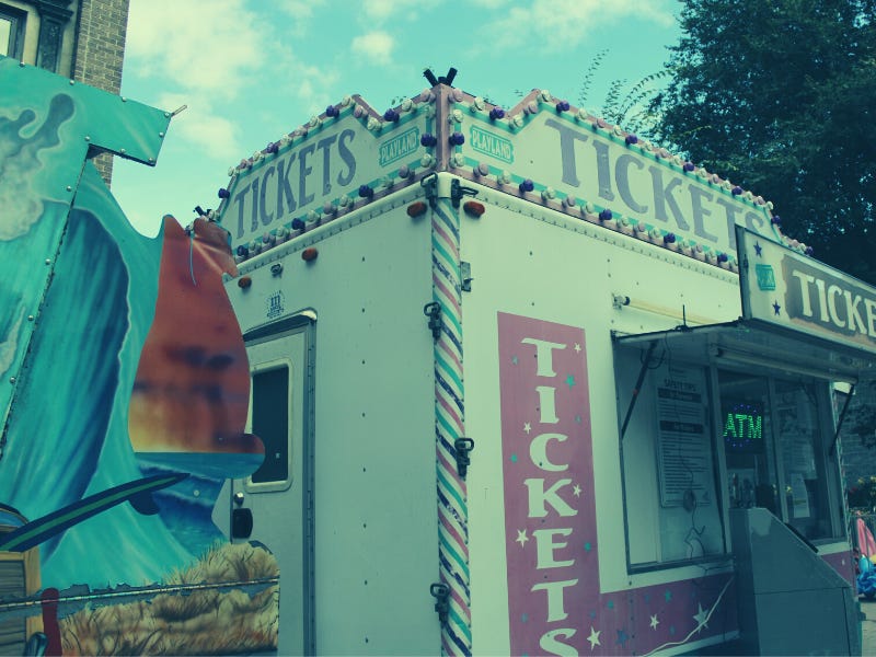 A faded photo of a carnival funhouse next to a portable ticket booth. The booth is white with pink and blue trim and says "Tickets". Behind both is a blue sky. A faded photo of a carnival funhouse next to a portable ticket booth. The booth is white with pink and blue trim and says "Tickets". Behind both is a blue sky.