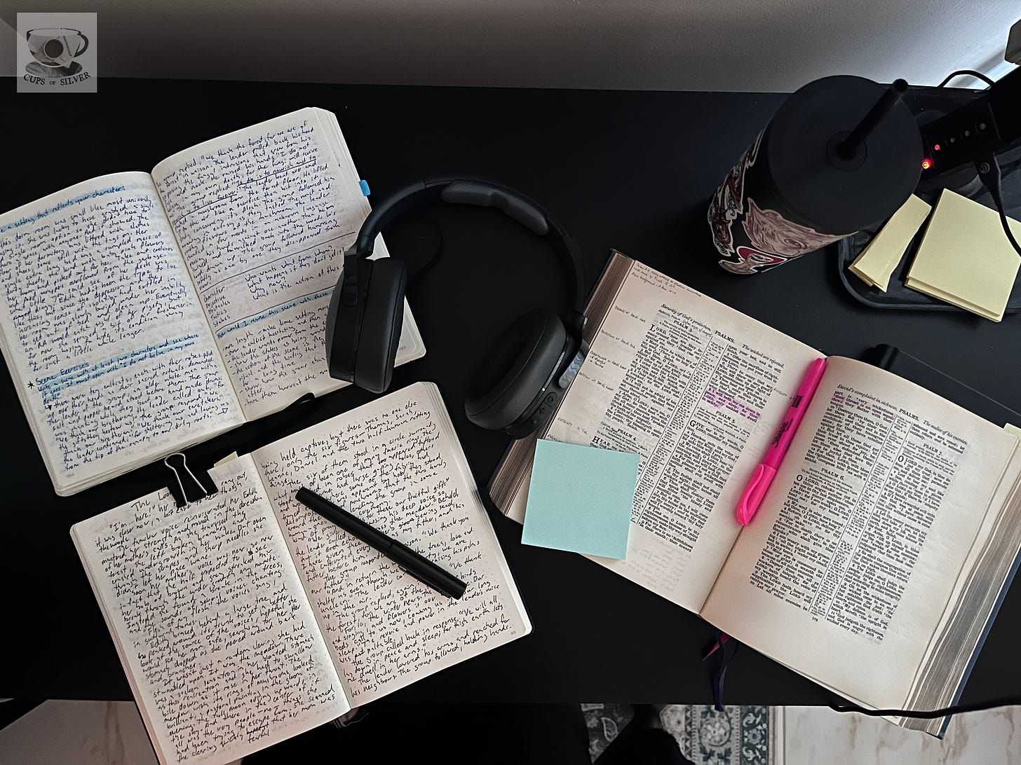 Photo of two journals open with handwriting in them, a pair of headphones, and the bible laid open on a black desk. Photo of two journals open with handwriting in them, a pair of headphones, and the bible laid open on a black desk.