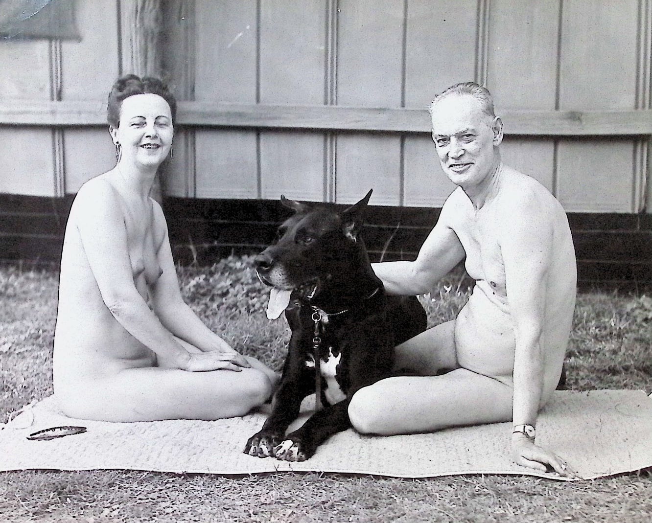 Black-and-white photograph of an older couple sitting nude on a woven mat on grass beside a wood-paneled building. A woman with dark upswept hair and hoop earrings sits at left; a balding man sits at right with his hand resting on a large black Great Dane lying between them, its tongue out.