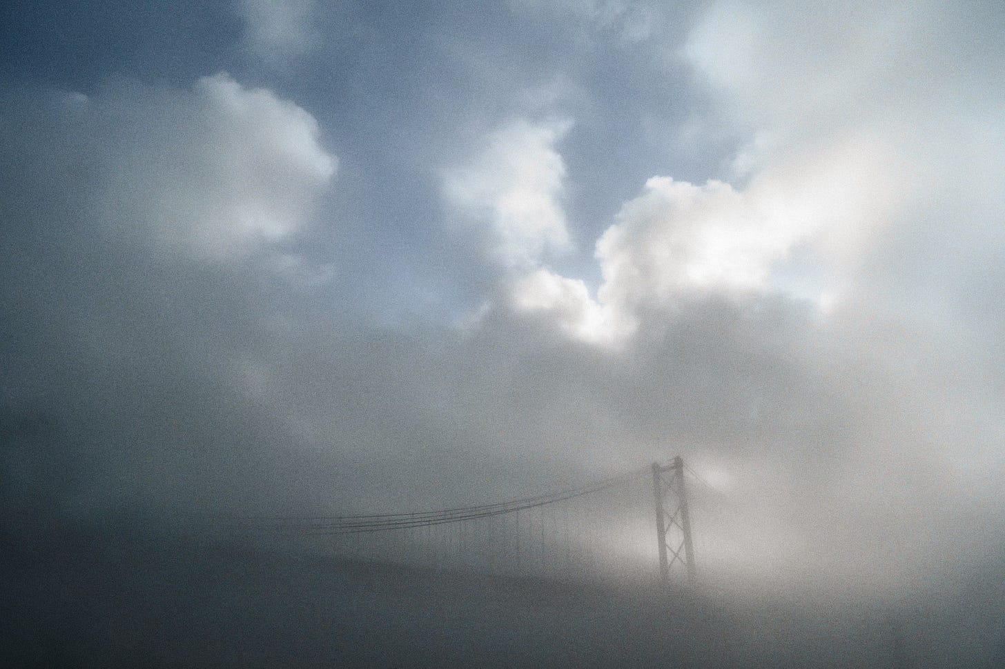 The 25 de Abril bridge is seen through fog and clouds, with blue sky on the top left side of the image