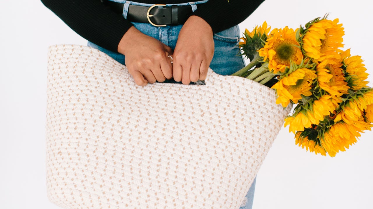 Hands gripping a textured woven bag with a bouquet of sunflowers peeking out. Photo by Social Curator.