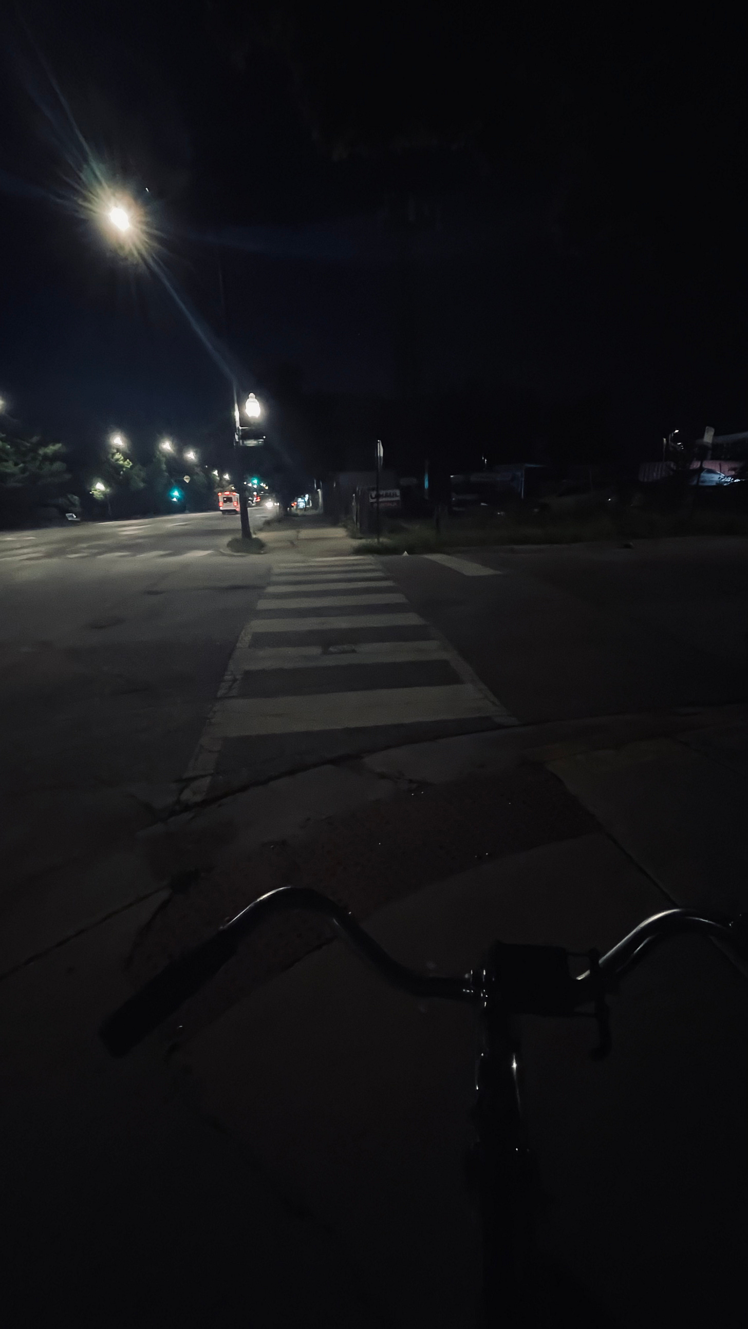 a street at night: the photo is taken while on a blue bicycle, with chrome handlebars, which glint in the dark. The corner of a block is ahead: a streetlight shines down onto the pavement.