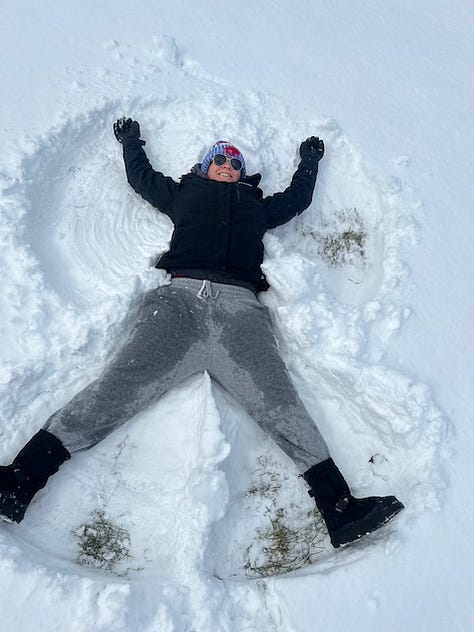 Woman makes a snow angel. Woman in a beanie cap, black jacket, and pink aviator glasses smiles in front of snow. A giant snow person hi-fives a lesbian with it's twig arm.