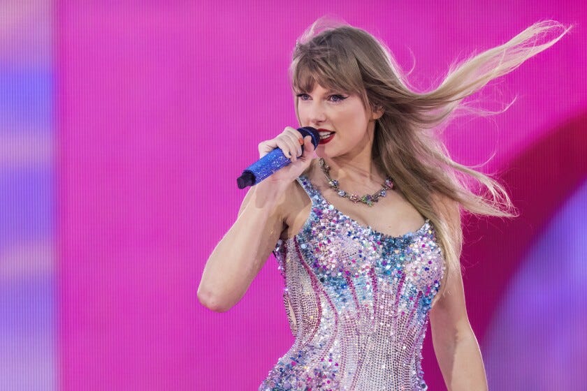 Taylor Swift, on a stage against a pink and purple backdrop and wearing a sequined costume, sings into a microphone.
