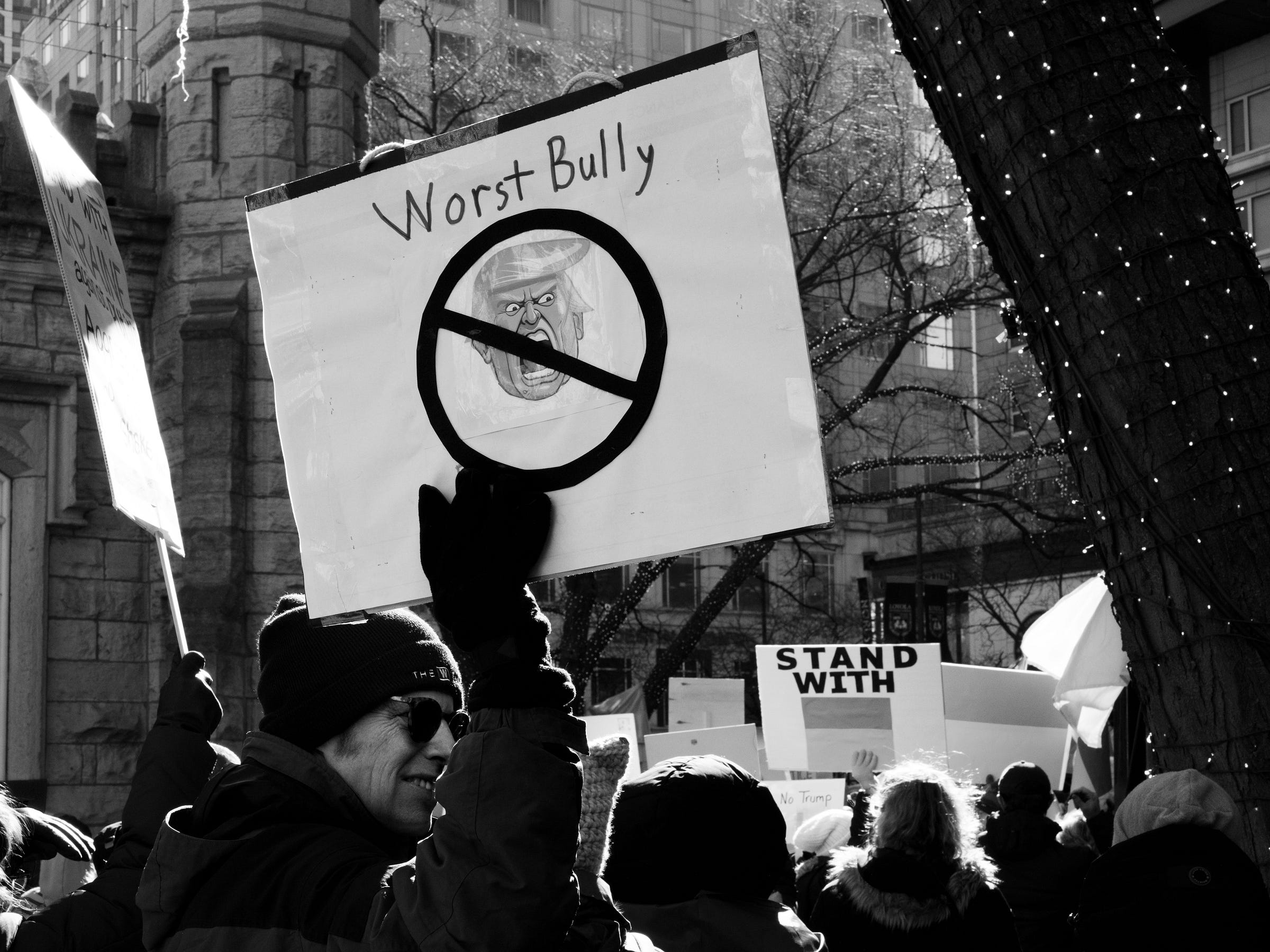 A black and white photograph of a protest captures a demonstrator holding a sign that reads "Worst Bully," featuring a caricature of Donald Trump with a crossed-out symbol over his face. The protest takes place in an urban setting, with a historic stone building and modern high-rises in the background. Other protesters carry signs, including one that reads "Stand With," partially obscured. A tree wrapped in string lights frames the right side of the image, contrasting the winter-clad crowd gathered in resistance. A black and white photograph of a protest captures a demonstrator holding a sign that reads "Worst Bully," featuring a caricature of Donald Trump with a crossed-out symbol over his face. The protest takes place in an urban setting, with a historic stone building and modern high-rises in the background. Other protesters carry signs, including one that reads "Stand With," partially obscured. A tree wrapped in string lights frames the right side of the image, contrasting the winter-clad crowd gathered in resistance.
