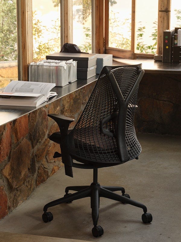 Herman Miller Sayl chair in black, viewed from behind, positioned at a stone-faced desk in a sun-lit studio with large windows and foliage outside.