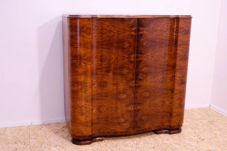 May include: A vintage wooden cabinet with two doors. The cabinet is made of dark wood with a rich grain pattern. The doors are slightly curved and have a simple design. The cabinet is standing on a light brown wooden floor.