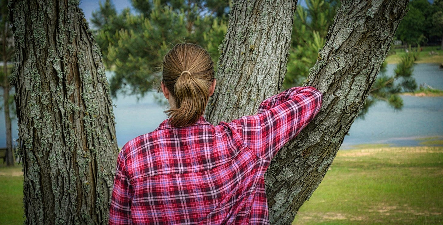 Young woman with a pony tail facing the lake in a red flannel shirt and leaning against a tree with blue sky and bits of lake in the background