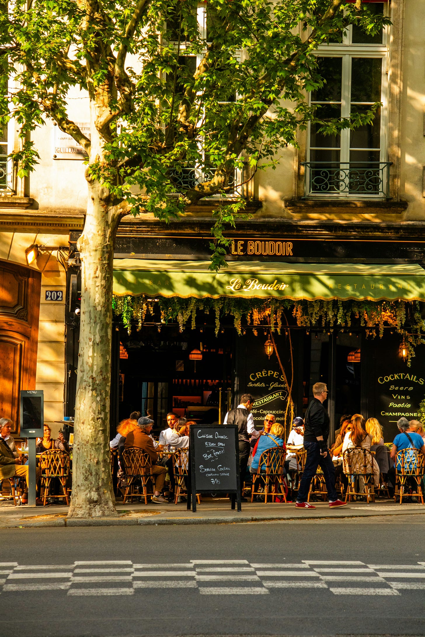 Parisian cafe with patrons sitting on tables by the sidewalk. The café is called "Le Boudoir"