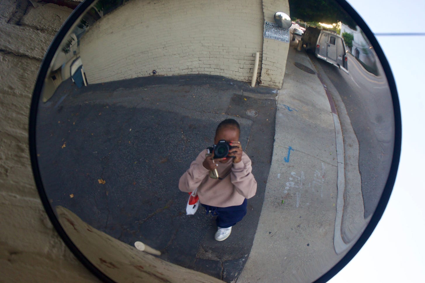 A photographer captures her reflection in a circular street mirror, her face hidden behind the camera lens as she stands alone in a quiet alley — a visual meditation on identity, distance, and becoming. A photographer captures her reflection in a circular street mirror, her face hidden behind the camera lens as she stands alone in a quiet alley — a visual meditation on identity, distance, and becoming.