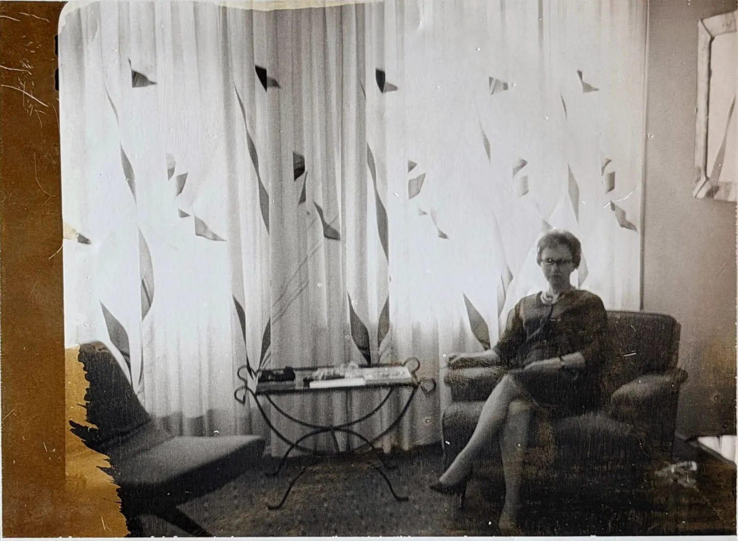 Photo ancienne en noir et blanc de Mamie Rose plus âgée, assise dans un fauteuil de salon devant des rideaux transparents, en robe et collier de perles.