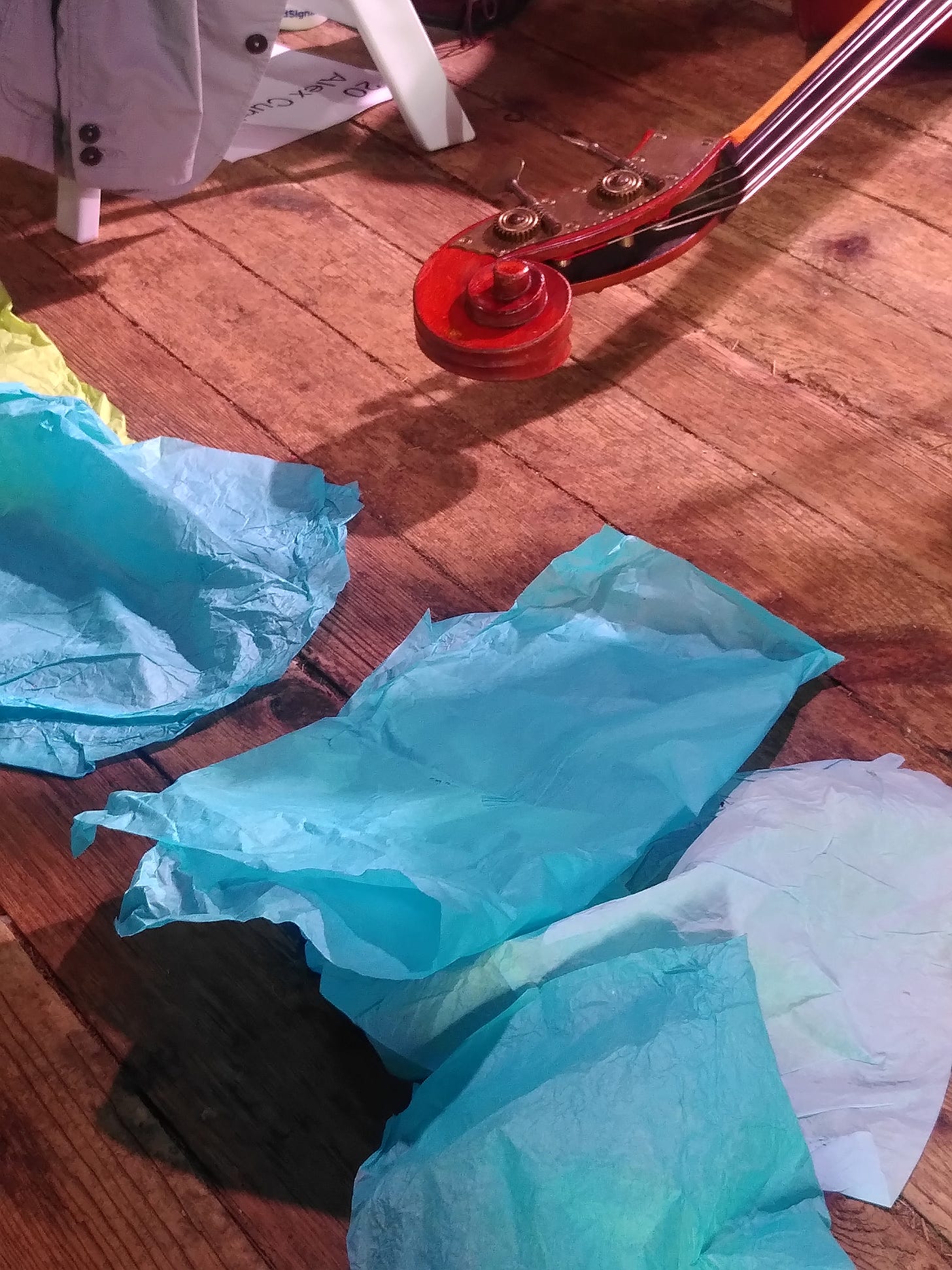 Bright blue tissue paper rests on hardwood floors while the head of cello peeks in the top right hand corner.