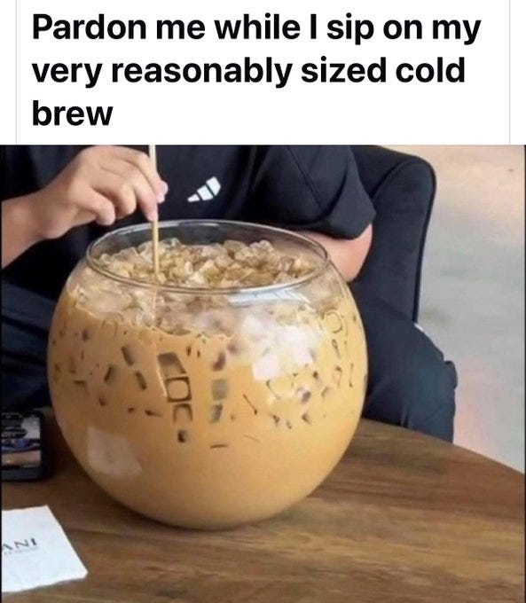 A person holding a straw, drinking from a large glass filled with iced cold brew coffee. The glass is transparent, containing ice cubes and a brown liquid, placed on a wooden table. The person is wearing a black shirt with white stripes on the sleeve.