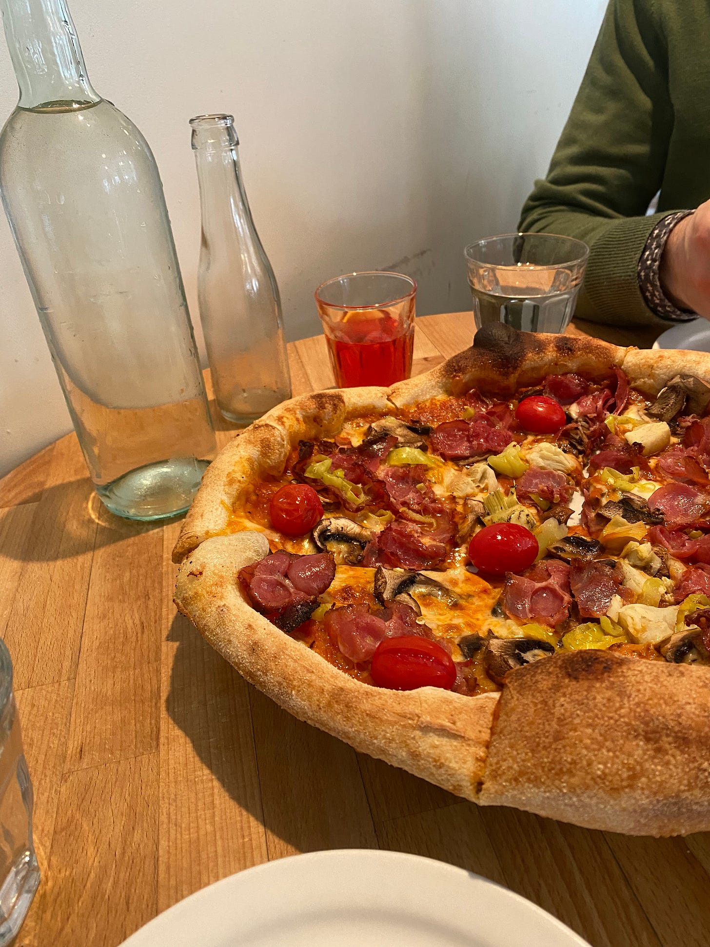 The pizza described above, with bright red tomatoes and vegetables and crisp meat, a puffed, browned crust. About 3/4 of it is in the frame on top of a wood table. Next to the pizza is a wine bottle full of water, and a small empty bottle with a little glass full of bicicletta cocktail. Jeff's arm, in a green sweater and a dark shirt cuff showing beneath it, is visible on the other side of the table.